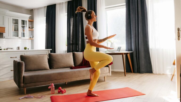 Full length of happy young sporty woman in headphones doing aerobics while training at home